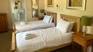 Een hotelkamer met twee eenpersoonsbedden, een houten nachtkastje, een nachttafeltje met een lamp, een spiegel, een badkamer met een wastafel en toilet, en een deur die naar een andere kamer leidt.