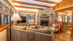 A cozy living room with a stone fireplace, wooden beams, and a large window. - Cedar Cottage by Hauserman Rentals (Homewood)