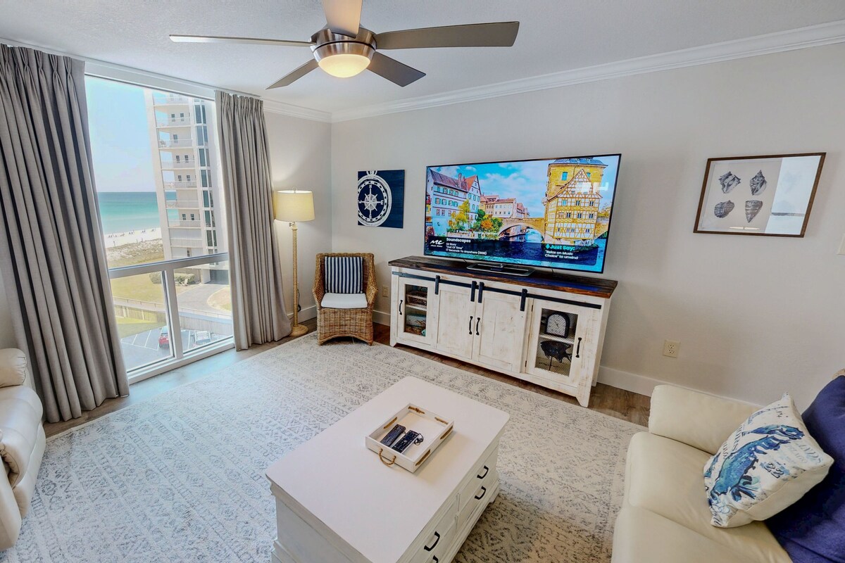 A hotel room with a large window, a ceiling fan, a flat-screen TV, a wicker chair, a white coffee table, and a sofa.