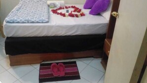 A bed with a patterned bedspread, a heart-shaped arrangement of red flowers, and two purple pillows. - NevaLand County Hotel (Matiliku)