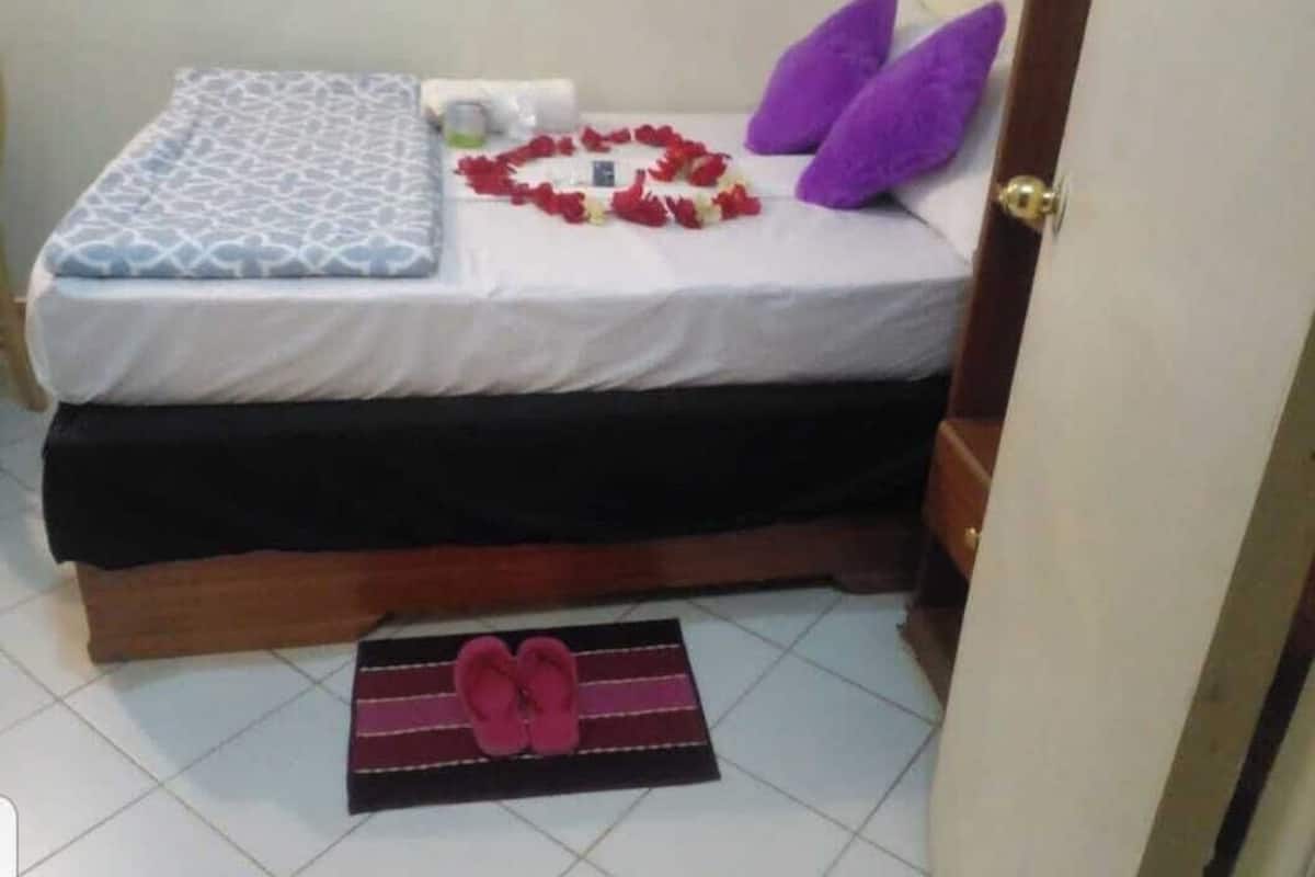 A bed with a patterned bedspread, a heart-shaped arrangement of red flowers, and two purple pillows.