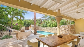 A covered outdoor area with a wooden table and chairs, a hammock, and a swimming pool. - Villa The Hidden Palm, Piscadera, Private Pool (Willemstad)