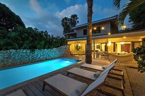 A pool area with lounge chairs, a stone wall, and a building with outdoor seating. - Villa The Hidden Palm, Piscadera, Private Pool (Willemstad)