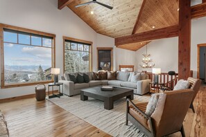 Ein geräumiges Wohnzimmer mit einer großen Eckcouch, einem Couchtisch und einem Kamin. Große Fenster bieten einen Blick auf die Berge.