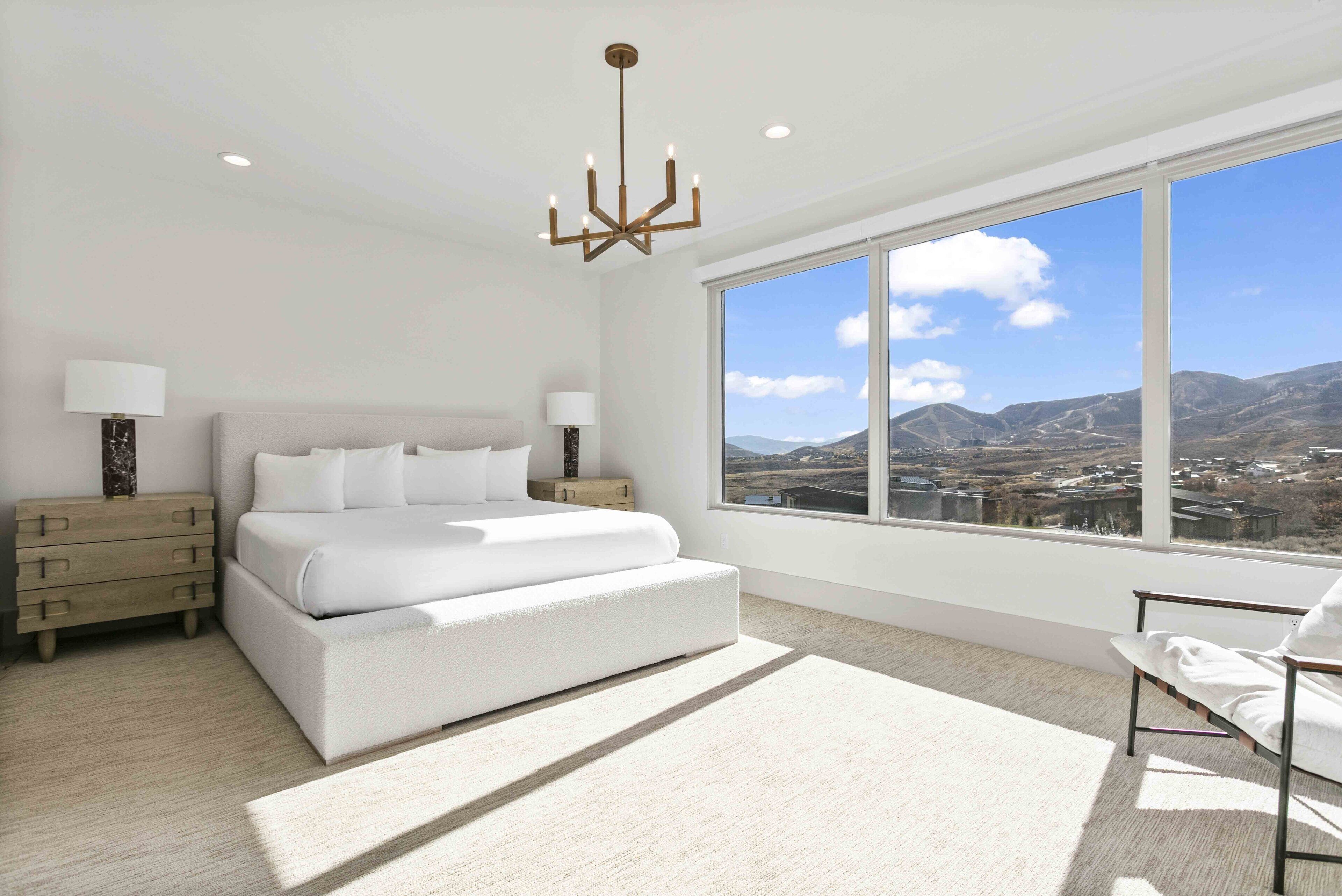 Une chambre moderne avec un grand lit, des tables de chevet, un lustre et une vue sur les montagnes par une grande fenêtre.