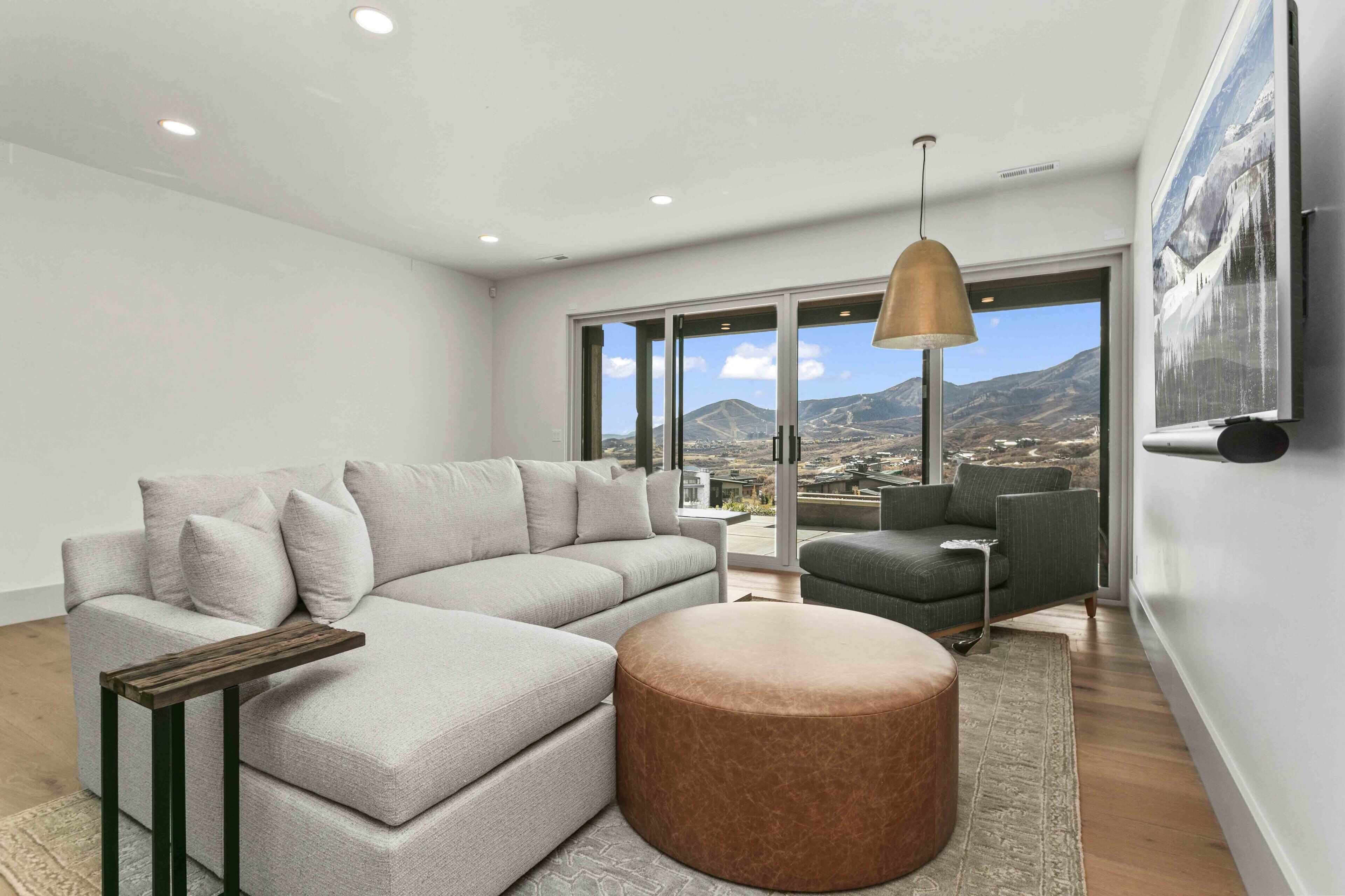 Una sala de estar moderna con un sofá seccional, un pouf redondo, una mesita auxiliar y un amplio ventanal con vistas a la montaña.