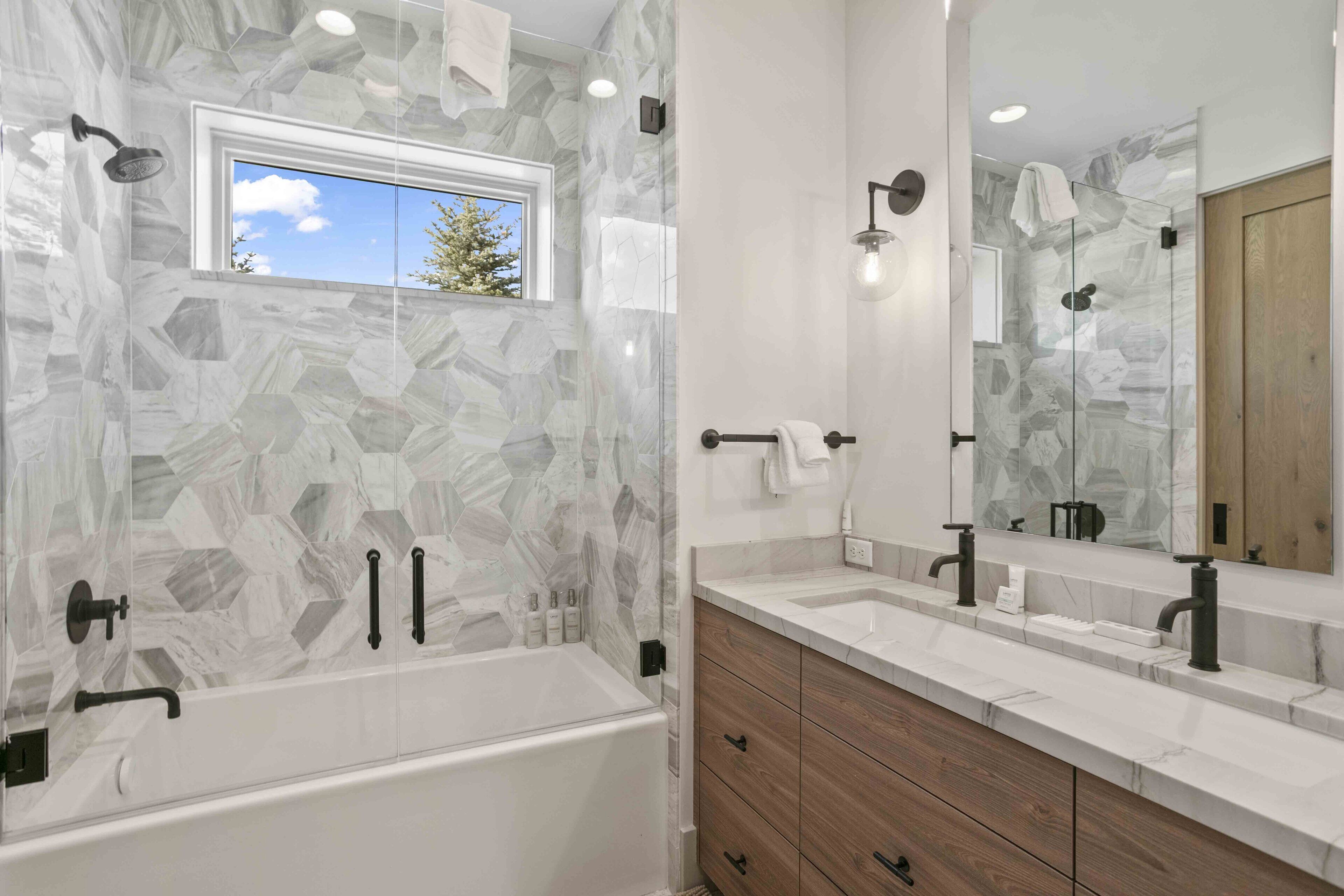 Un baño moderno con pared de diseño de piedra, una ventana, una bañera, un tocador con espejo y zona de ducha.