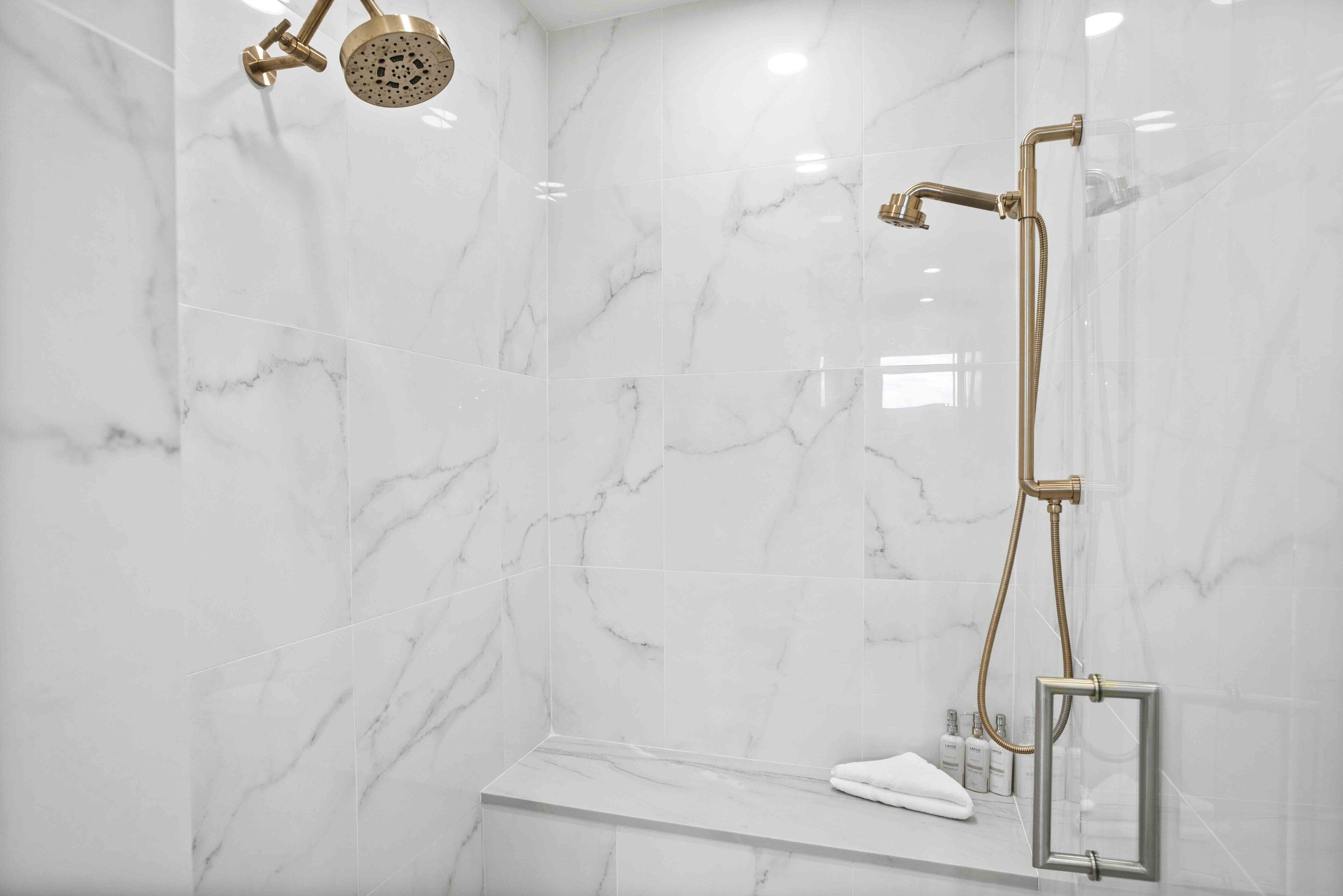 Une salle de bain moderne avec un mur en marbre, une douche dotée d’une robinetterie dorée et un banc où sont posés les articles de toilette.