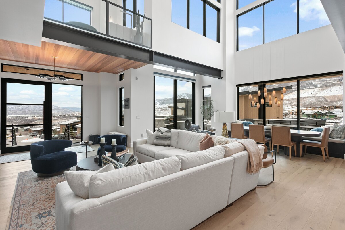 A spacious living area with a large white sectional sofa, a dark blue armchair, and a dining table with chairs. Large windows offer views of a snowy landscape.