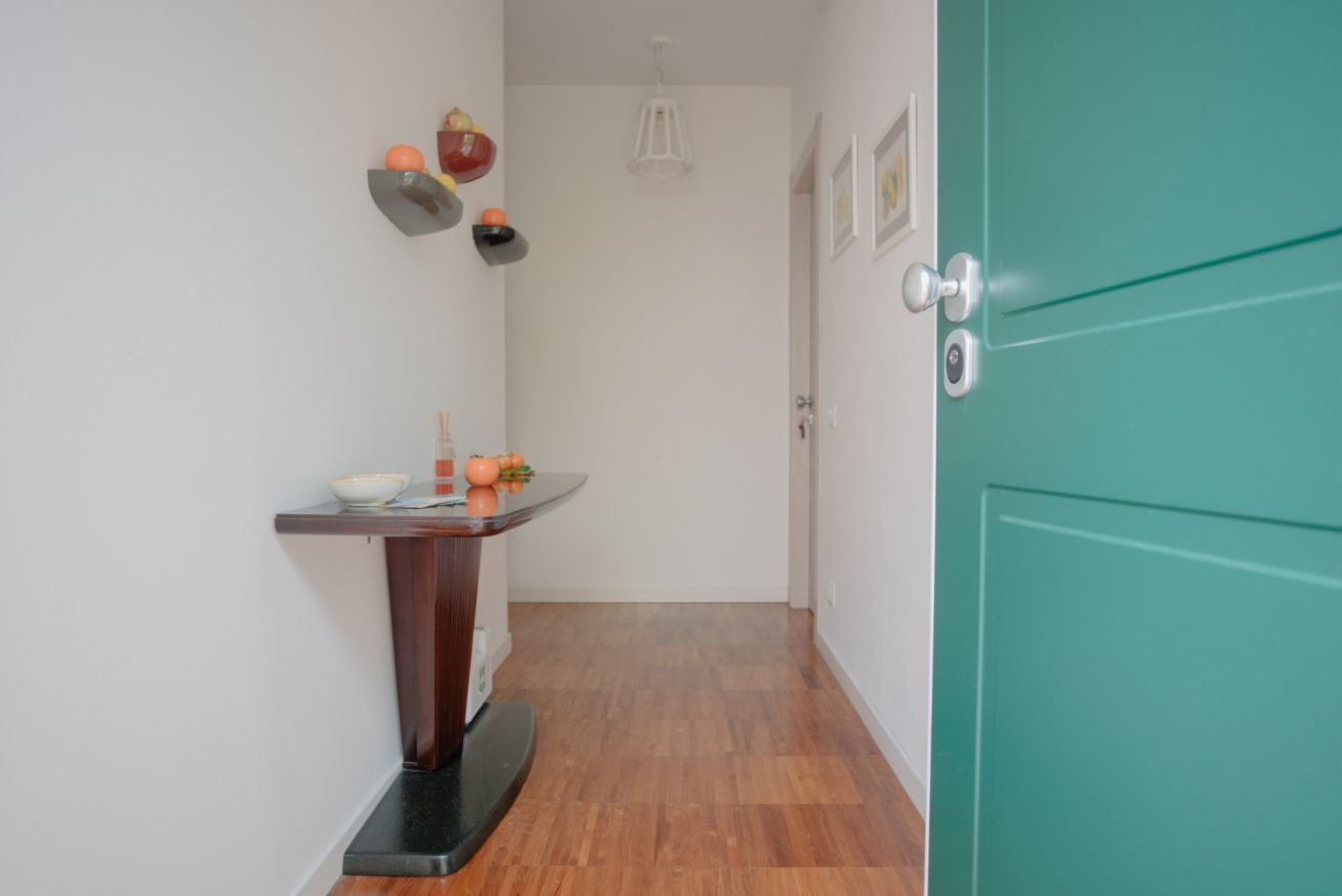Un pasillo estrecho con suelo de madera, una puerta color esmeralda a la derecha y una pequeña mesa con frutas y un cuenco a la izquierda.
