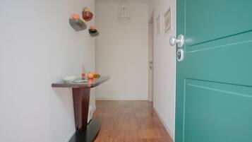 Un pasillo estrecho con suelo de madera, una puerta color esmeralda a la derecha y una pequeña mesa con frutas y un cuenco a la izquierda.