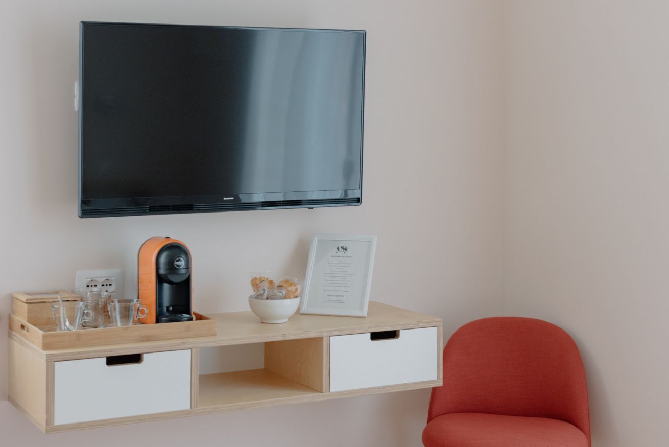 Una habitación moderna con una televisión de pantalla plana montada en la pared, una mesita de madera con una cafetera, vasos y un menú enmarcado, y una silla roja.