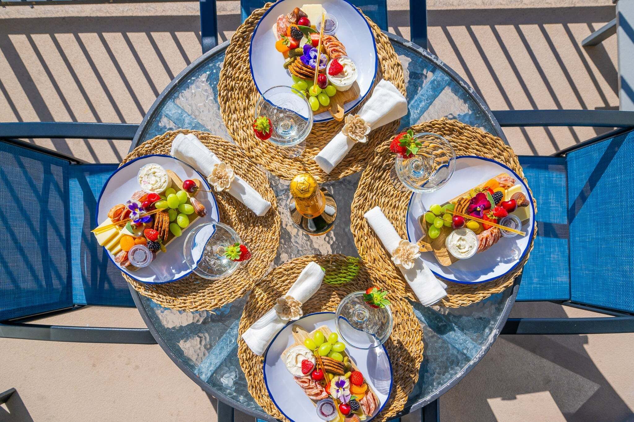 Una mesa redonda al aire libre con cuatro platos de frutas y quesos variados, cada uno con un vaso de agua y una servilleta.