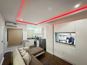 A modern living room with a sofa, a TV, and a kitchenette in the background.