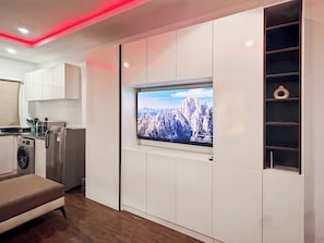 A modern kitchen with a built-in washing machine, a flat-screen TV mounted on a white cabinet, and a wooden bench.