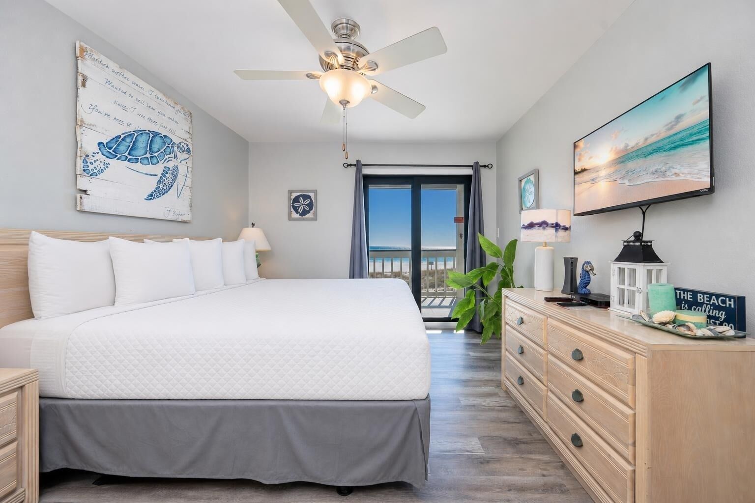 A bedroom with a bed, a ceiling fan, a dresser, a TV, and a view of the beach.