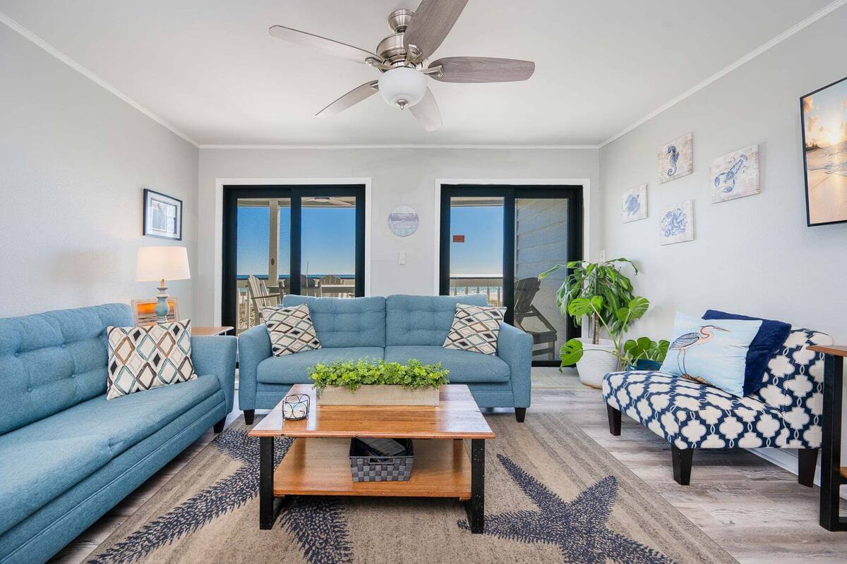 A living room with a blue sofa, a wooden coffee table, and a patterned armchair. There is a ceiling fan, a large window, and a view of the ocean.