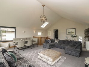 A spacious living room with a high ceiling, a dining area with a wooden table and chairs, and a cozy seating area with a patterned sofa and ottoman. - Two Hollens (Ambleside)