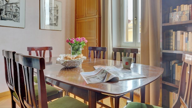 Un comedor con mesa de madera, sillas, un florero con flores y una estantería al fondo.