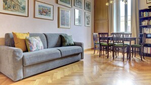 Una sala de estar con un sofá gris, cojines verdes y amarillos, una mesa de comedor de madera con sillas y una estantería llena de libros.