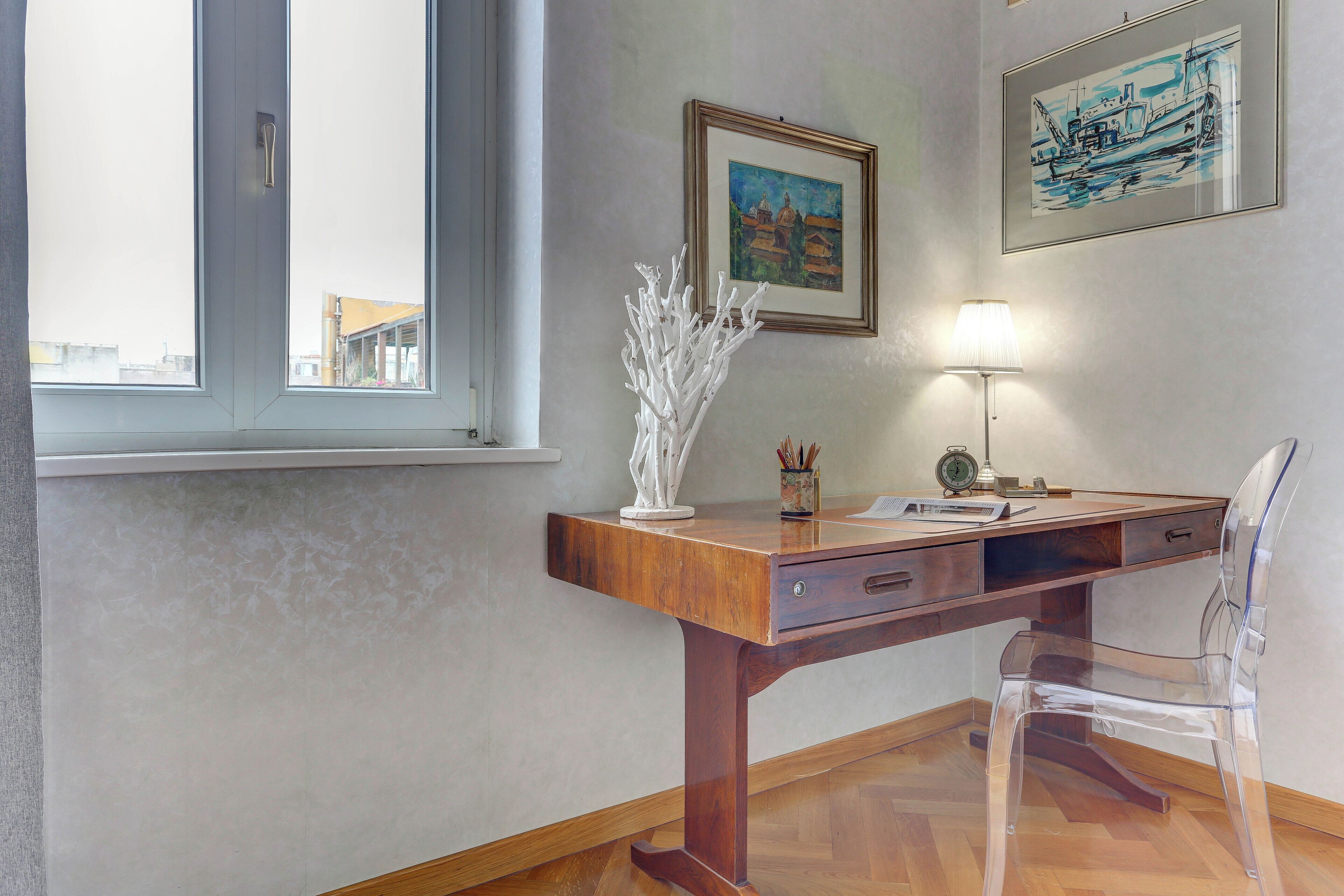 A wooden desk with a clear plastic chair, a lamp, and a framed painting on the wall.