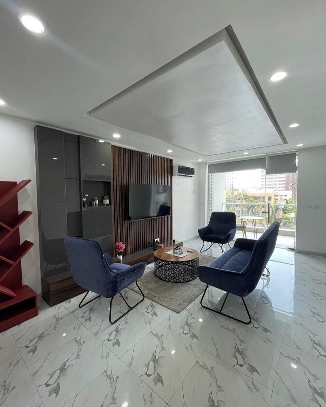 A modern living room with a flat-screen TV, two blue armchairs, a coffee table, and a red staircase.