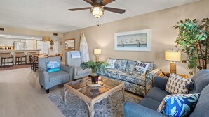 A living room with a ceiling fan, a sofa with beach-themed cushions, a coffee table, and a dining area with a kitchen in the background. - Cozy Beach Retreat Close to Beach w/Courtyard Pool, Kiddie Pool, Jacuzzi & Grills! (South Padre Island)