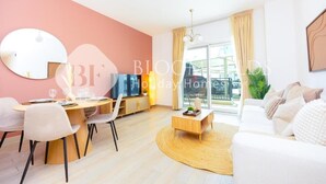 A modern living room with a round dining table, white sofa, and a large window with curtains. - Bloomfields Royal 2Br Water edge (Abu Dhabi)