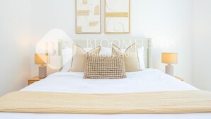 A neatly made bed with a beige blanket and two pillows, two wall-mounted abstract paintings, and two bedside lamps on a white nightstand. - Bloomfields Royal 2Br Water edge (Abu Dhabi)