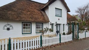 Une maison blanche traditionnelle, avec un toit de chaume, une clôture blanche en bois et un chemin de briques menant à l’entrée.