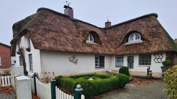 Une maison traditionnelle à toit de chaume, avec une façade blanche, une clôture en bois peinte en blanc et un jardin bien entretenu.