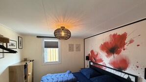 Et soveværelse med seng, et vindue, en radiator, en hylde med genstande og en væg med et stort abstrakt maleri.