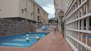 A swimming pool with a tiled deck, surrounded by buildings with balconies and air conditioning units.