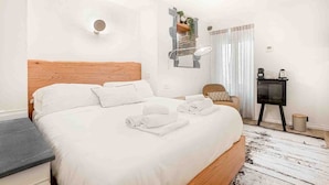Un dormitorio ordenado con cama de cabecera de madera, ropa de cama blanca, una mesita de noche, una silla pequeña, una mesita auxiliar y un estante en la pared con plantas, además de una ventana con cortinas.