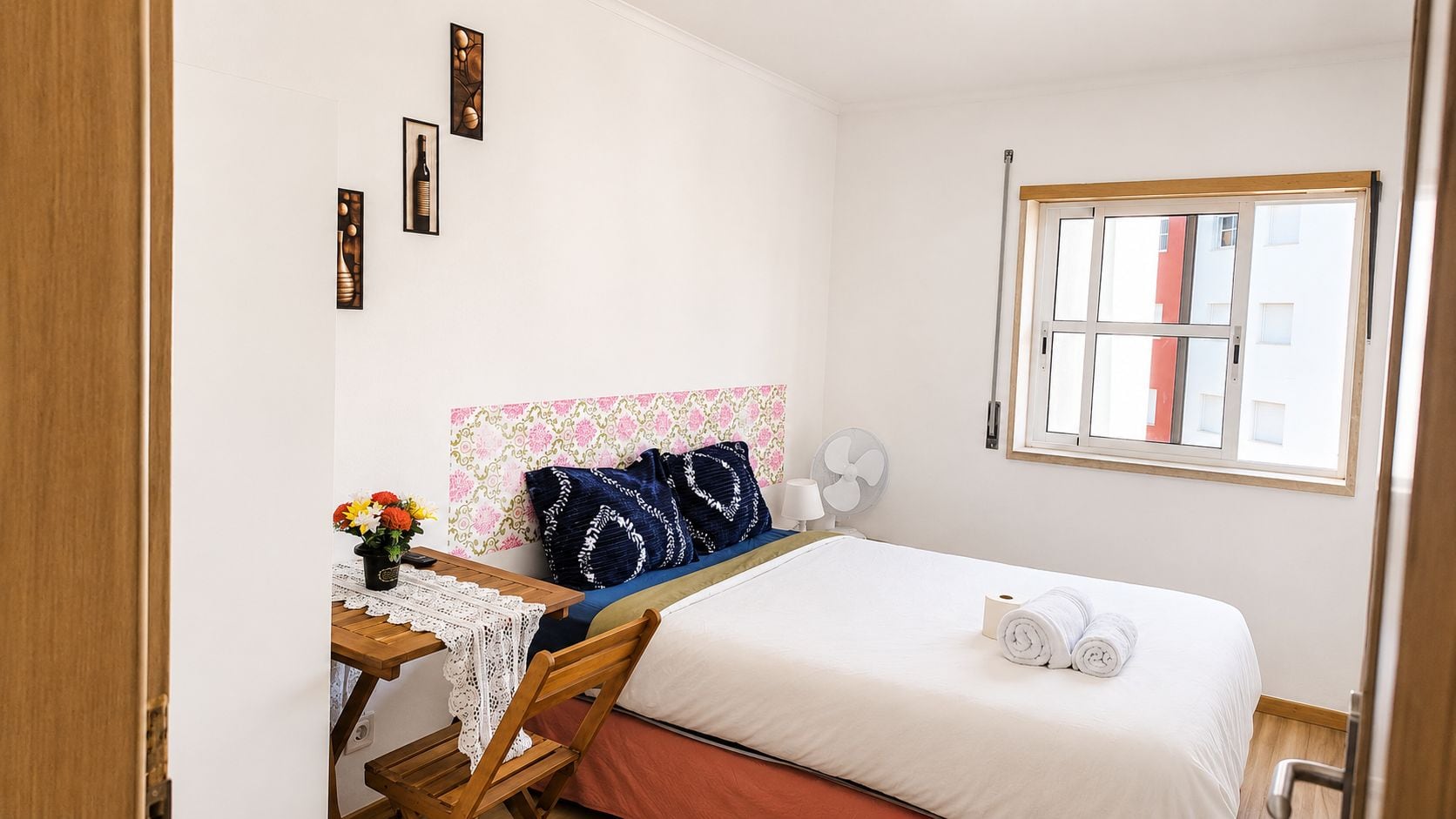 Un dormitorio con cama, un escritorio con silla, una ventana y un ventilador fijado a la pared.