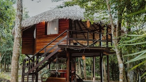 A wooden cabin with a thatched roof, surrounded by trees. It features a wooden staircase leading to a deck with a table and chairs. - Villa Arapiuns - 6 Bangalôs na Floresta (Santarém)