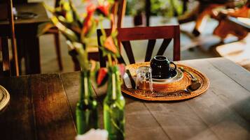 Uma mesa de madeira com um apoio de tecido trançado, uma chávena preta, um frasco de vidro e uma garrafa de água.
