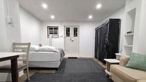Una habitación pequeña con cama, sillón, mesita, sofá y cortina negra.