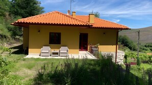 Uma casa amarela com telhado de telhas vermelhas, duas cadeiras brancas e uma pequena mesa na parte externa.