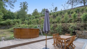 Uma área externa com mesa e cadeiras de madeira, uma banheira de hidromassagem rústica e um guarda-sol coberto.