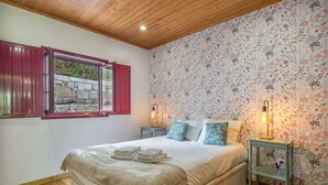 Quarto com cama, janela com portadas vermelhas, papel de parede floral e teto de madeira.