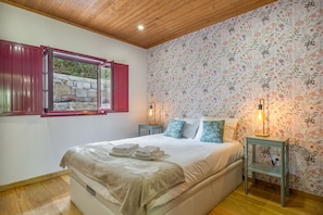A bedroom with a bed, a window with red shutters, floral wallpaper, and a wooden ceiling. - Casa dos Mirtilos, Germil – Mountain Retreat in Gerês (Germil)