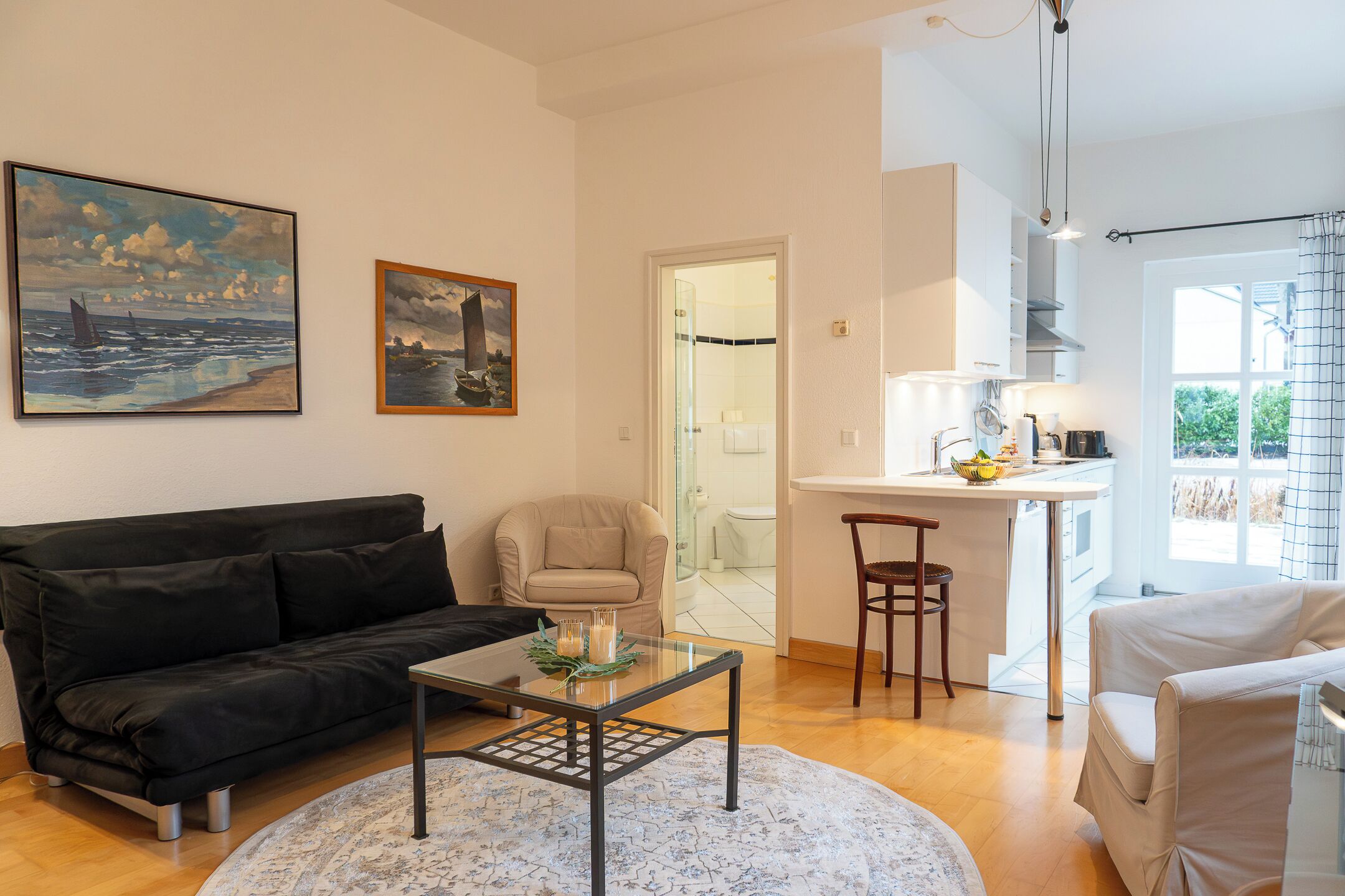 A living room with a black sofa, a glass coffee table, and a beige armchair. There is a kitchen area with a dining table and chairs, and a window with a view of greenery.