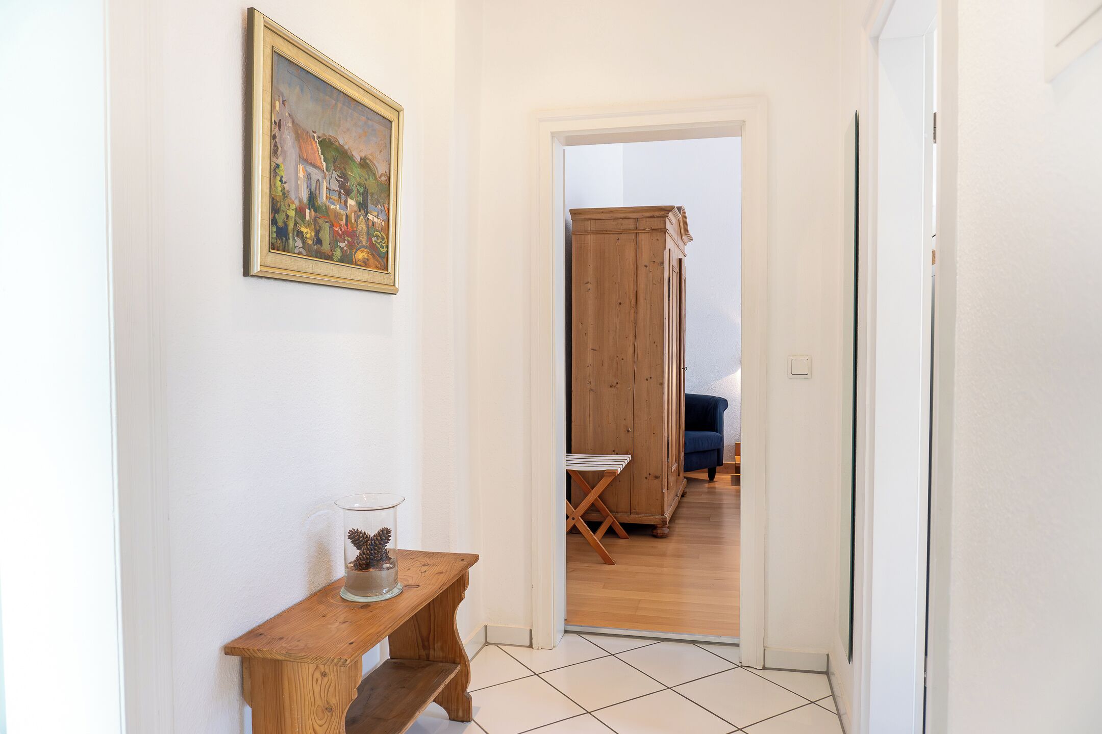A hallway with a wooden bench, a glass container with pinecones, and a painting on the wall.