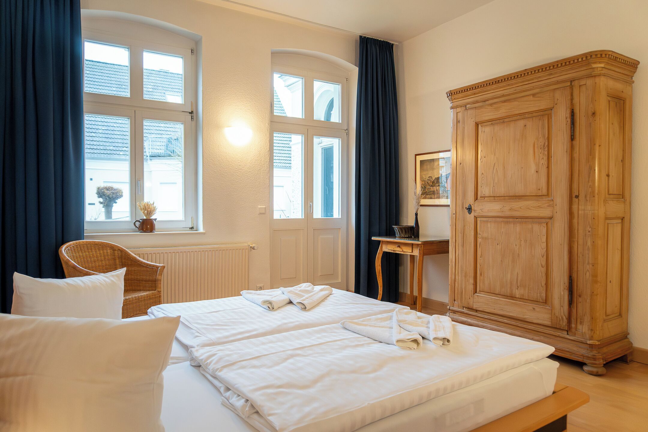 Ein Hotelzimmer mit zwei Betten, einem großen Holzschrank, einem Korbstuhl und einem kleinen Tisch mit einer Vase.