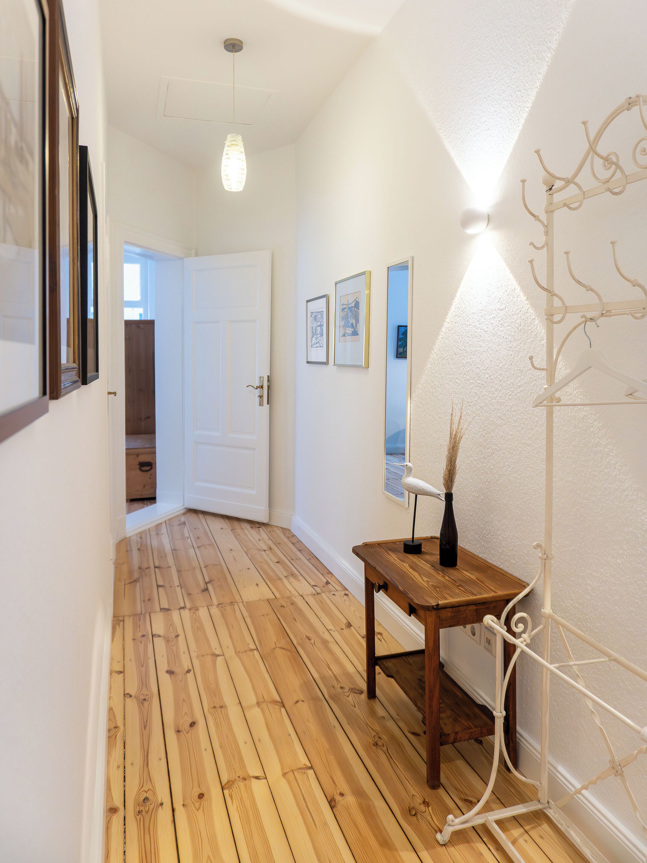 Un couloir doté d’un plancher en bois, d’une porte blanche et d’un porte-manteau sur lequel est posé un vase.