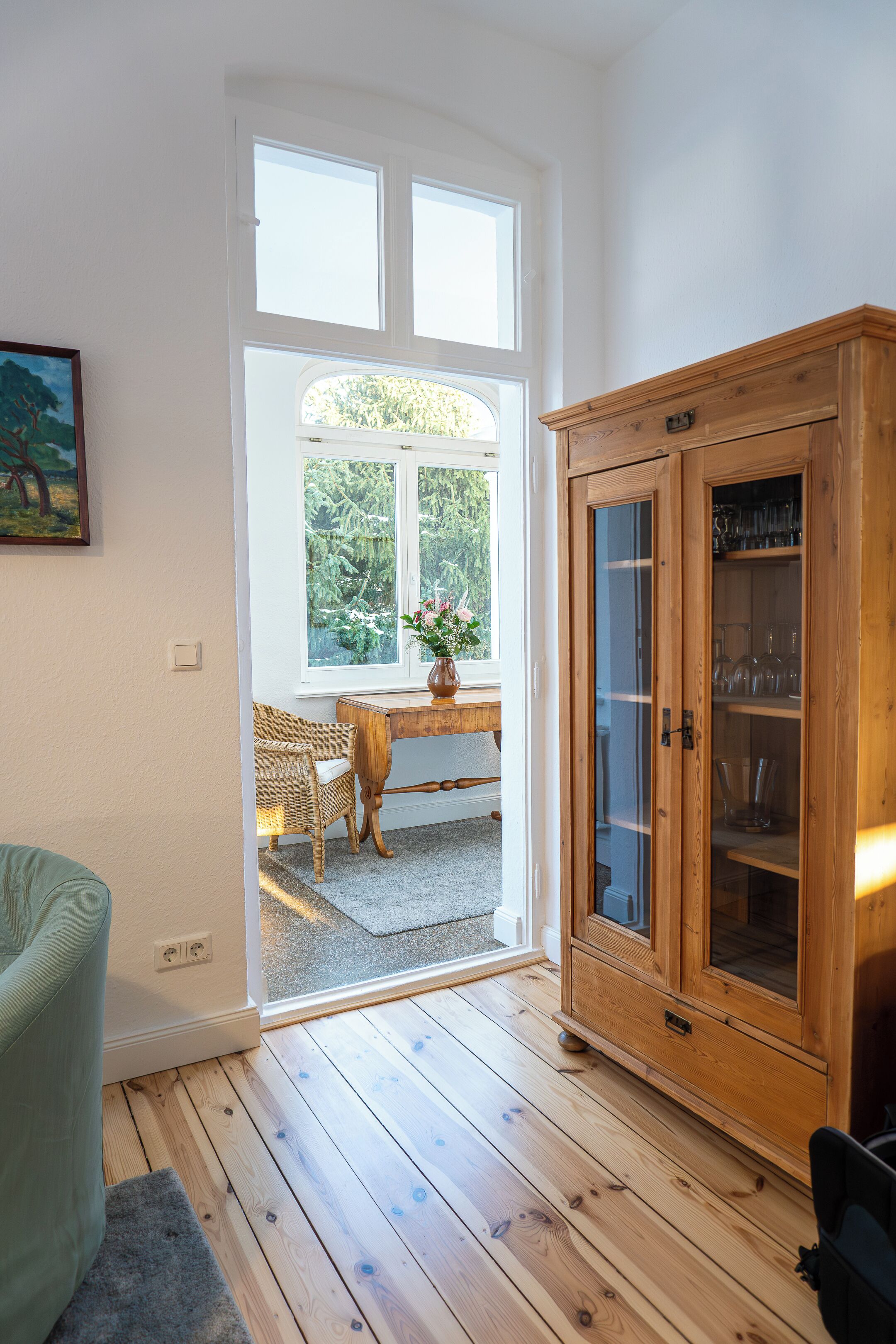 A room with wooden flooring, a green armchair, a wooden cabinet with glass doors, and a window with a view of greenery.