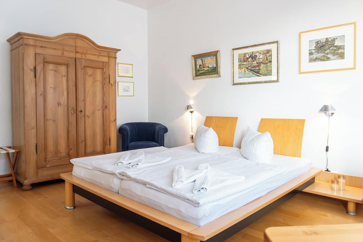A hotel room with a large wooden wardrobe, a bed with white linens, two bedside lamps, and three framed artworks on the wall.