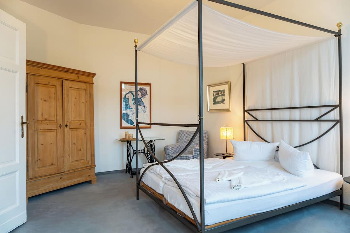 A four-poster bed with a canopy, a wooden wardrobe, a small table with a lamp, and a framed picture on the wall.