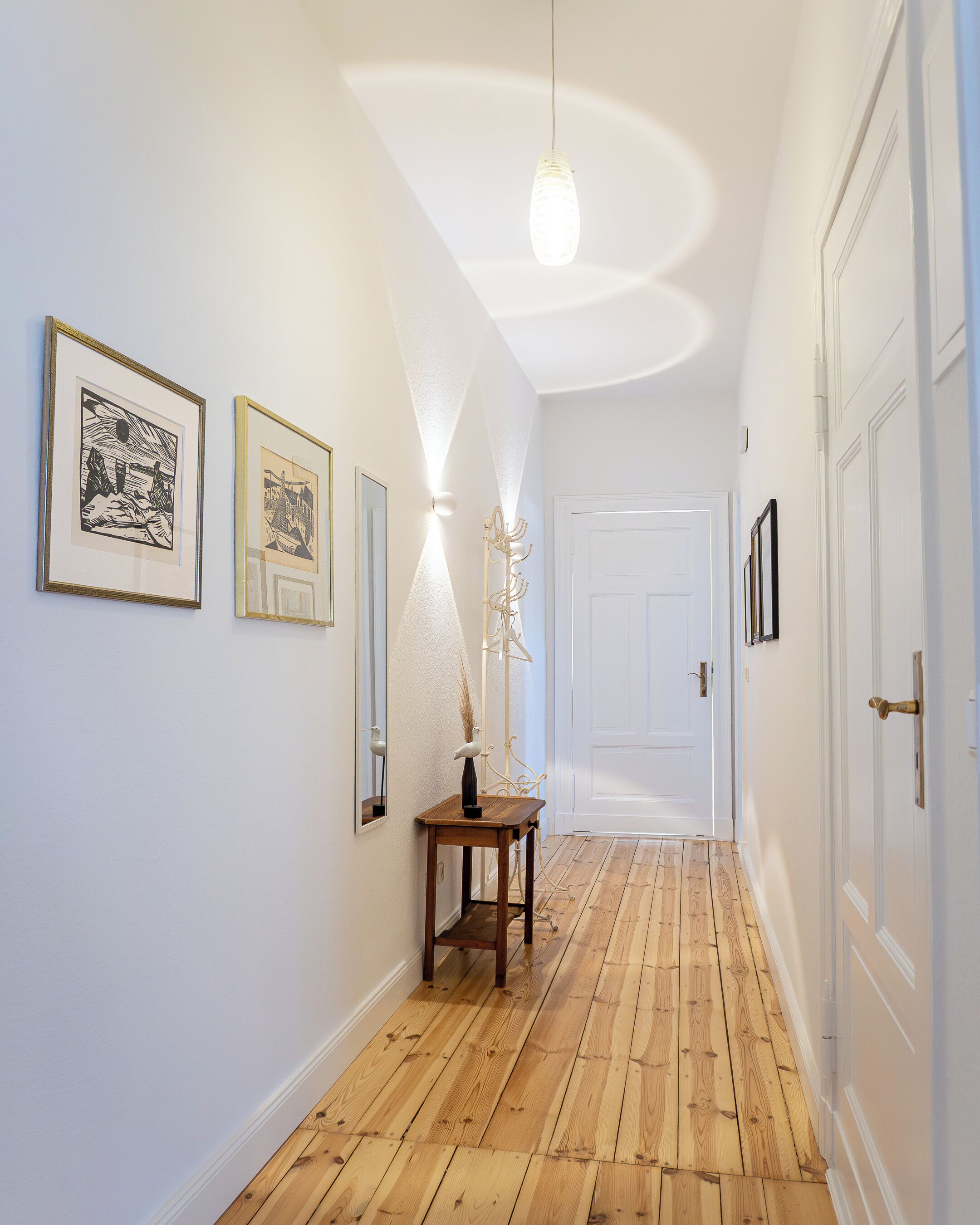 Un couloir doté d’un plancher en bois, de murs blancs et d’œuvres d’art encadrées. Une porte se trouve au bout du couloir, et un luminaire est suspendu au plafond.